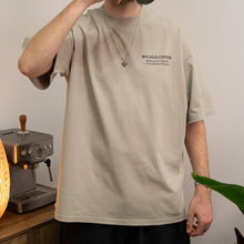 Cargar imagen en el visor de la galería, Person wearing a beige t-shirt with 'Big Dog Coffee Speciality Coffee for Everyday People', standing in a kitchen.
